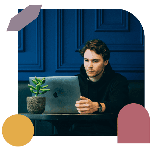 A person with brown hair sits at a table, focused on a laptop, with a small potted plant beside them. The background features a deep blue wall with decorative panels and colourful abstract shapes frame the image.