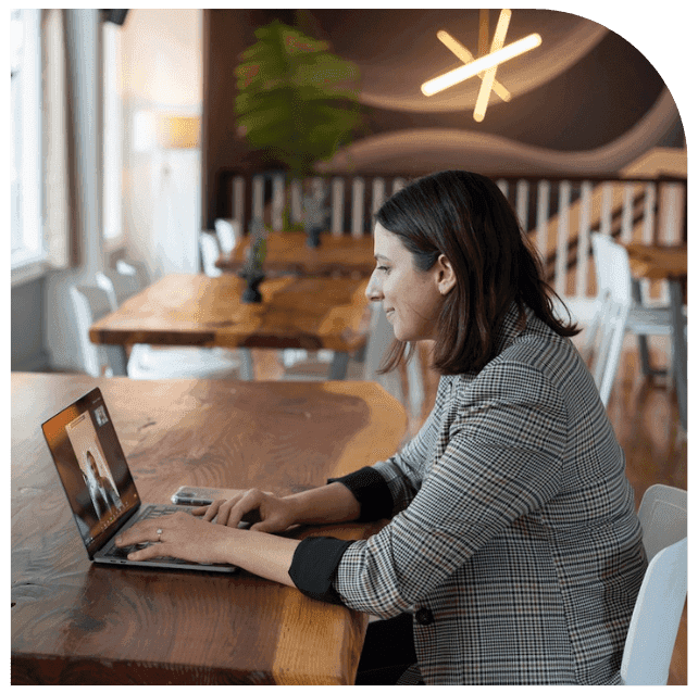 A woman in a checked blazer works at a laptop on a long wooden table in an empty cafe, a video call open on her screen.