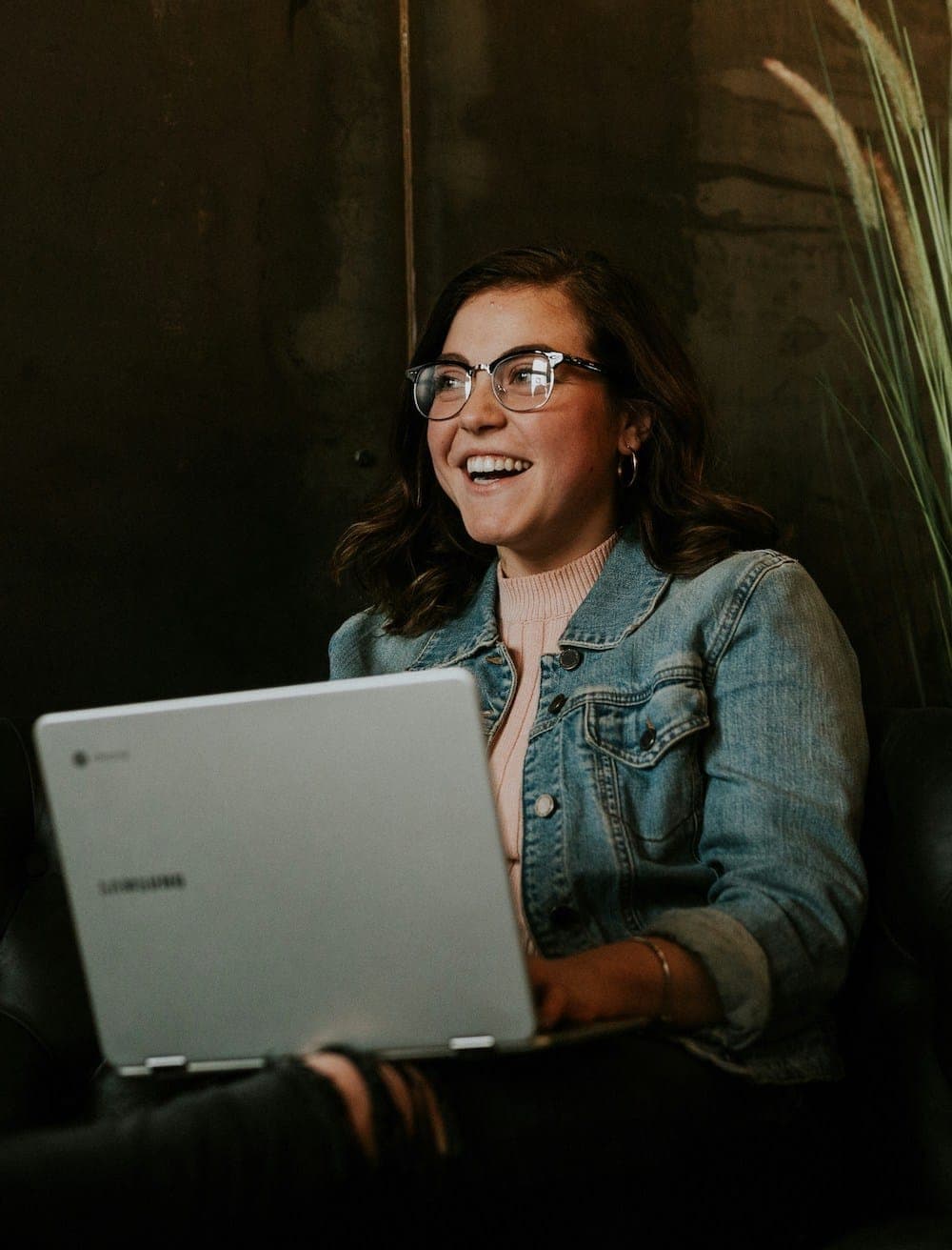 A woman in a denim jacket and glasses sits with a laptop on her lap, laughing as she looks off to one side.
