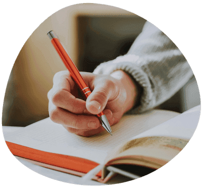 A close-up of a hand writing in an open ruled notebook with an orange pen.