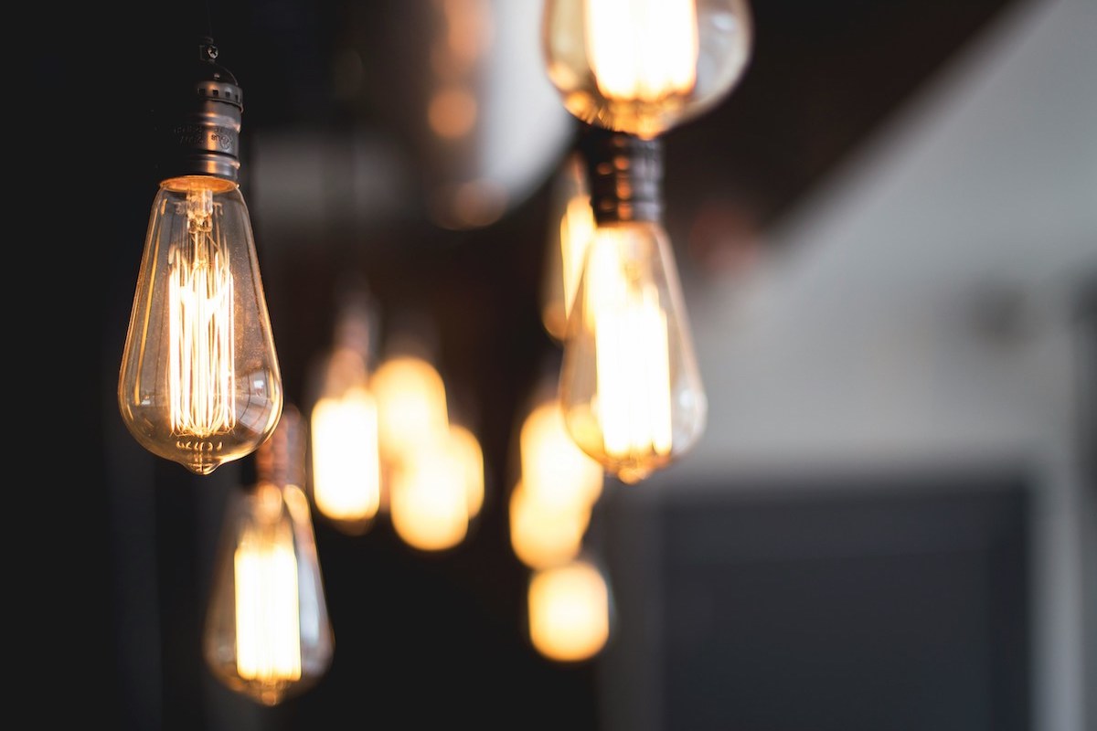 A row of vintage-style Edison light bulbs hanging from the ceiling, glowing warmly against a blurred dark background.