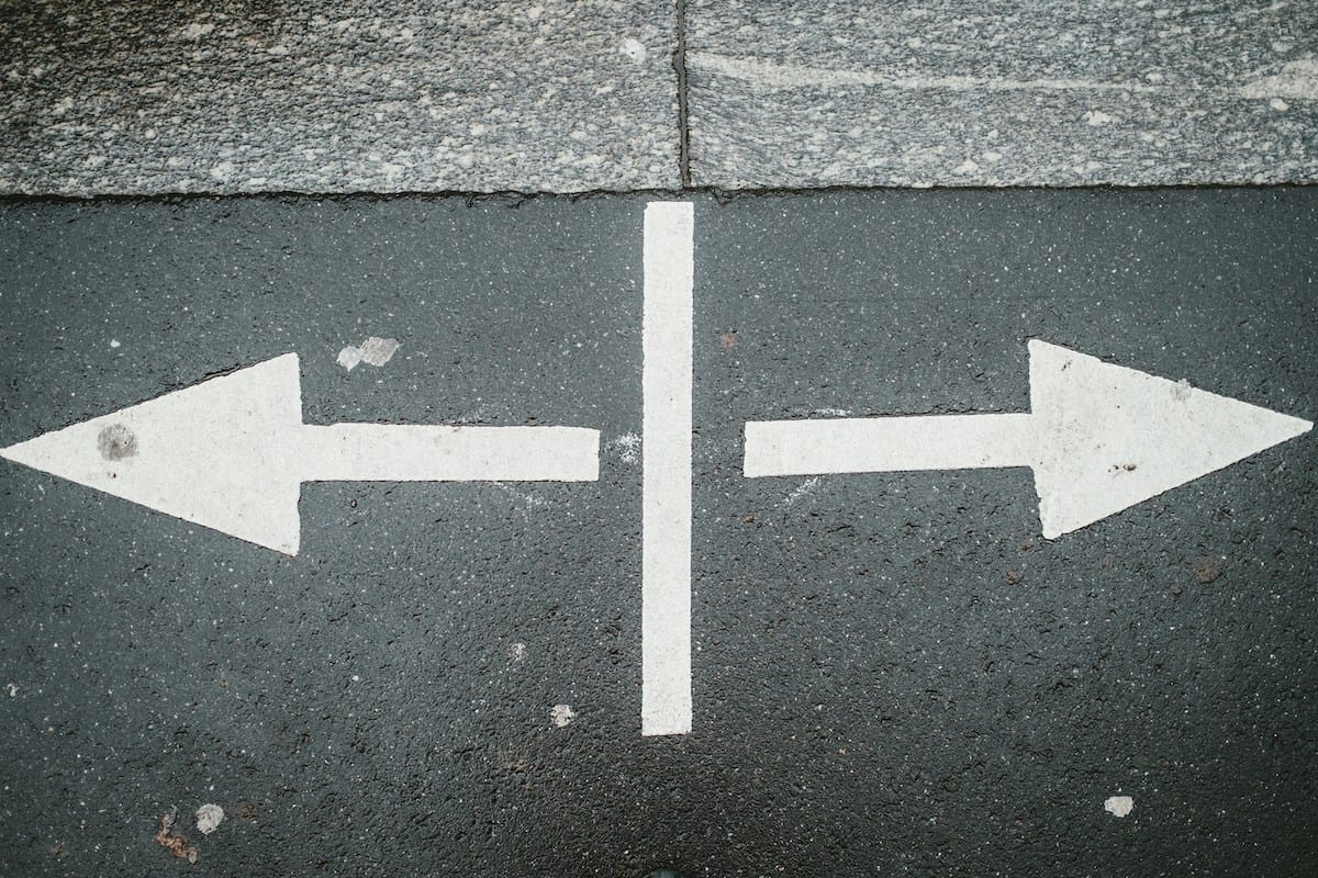 White painted arrows on asphalt point left and right, separated by a vertical white line. The surface appears slightly worn with visible texture.