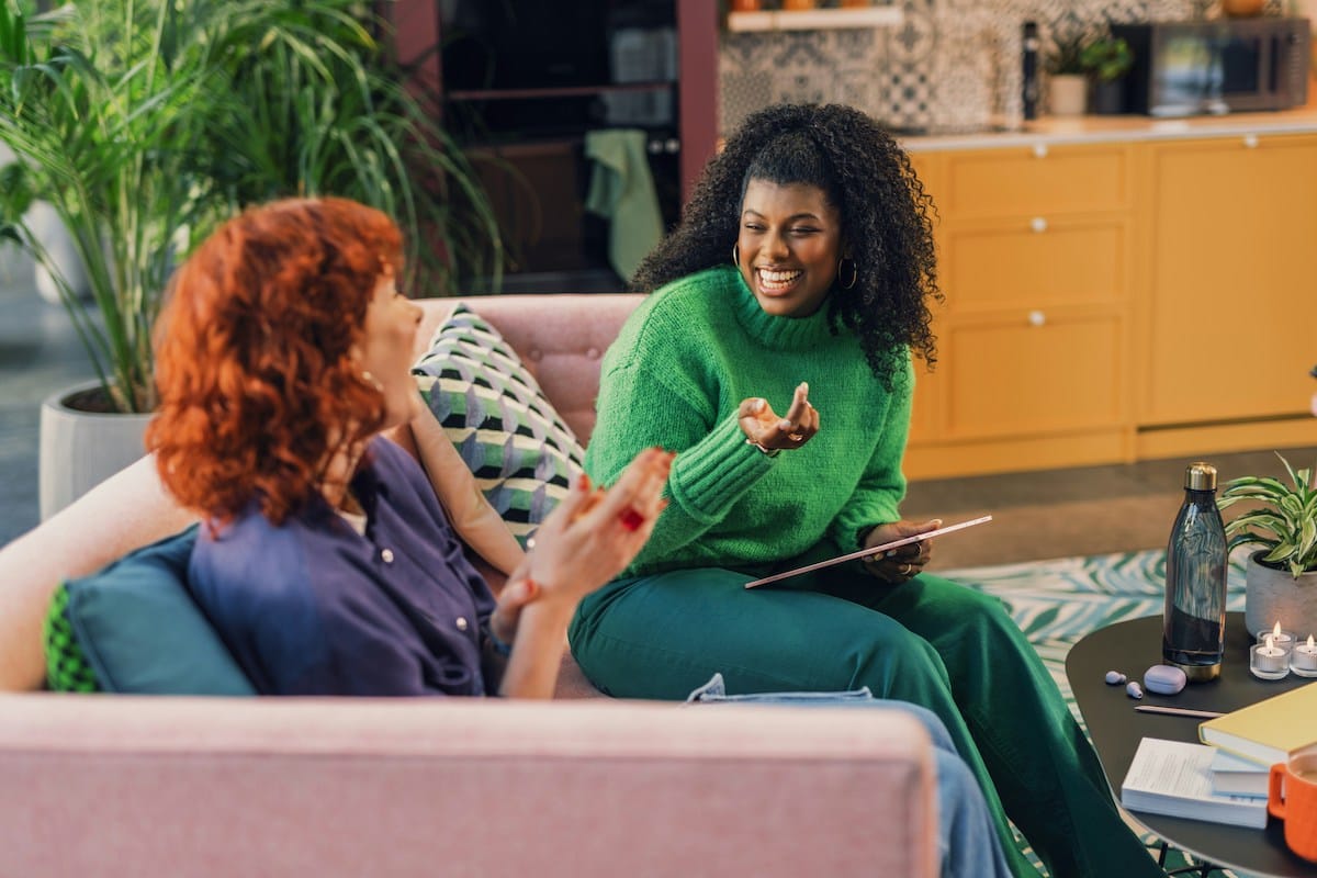 Two women sit on a pink couch in a cozy room, laughing and chatting. One wears a green sweater and holds a tablet, creating a joyful atmosphere.