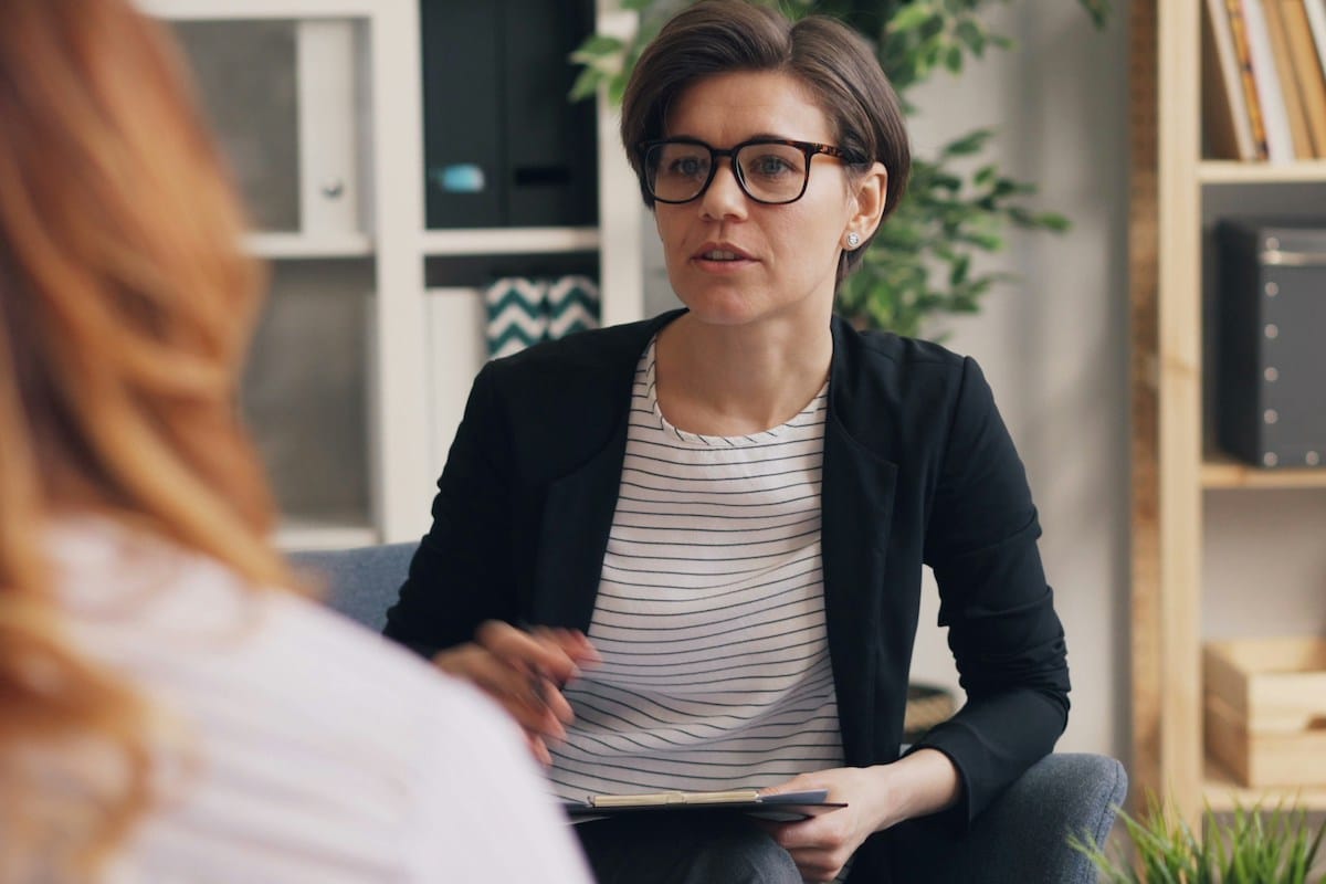 A woman with short hair and glasses, holding a clipboard, sits and talks to another person in an office setting.