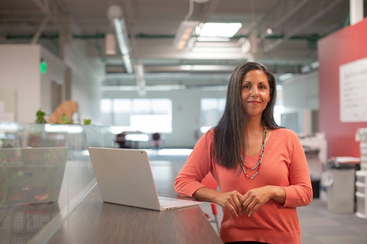 A woman stands in a modern office next to a laptop on a desk, looking at the camera and smiling slightly.
