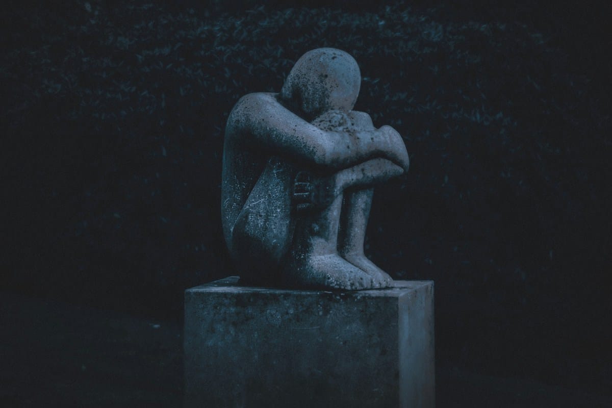 A concrete statue of a person sitting with knees drawn to chest and head resting on arms, displayed on a rectangular pedestal in a dimly lit outdoor setting.