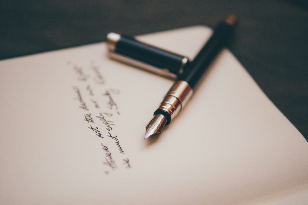 Elegant fountain pen with a black barrel and gold nib resting on cream-coloured paper, with handwritten cursive text visible but slightly blurred. The shallow depth of field and warm tones create a reflective, contemplative atmosphere evocative of journaling and self-expression.