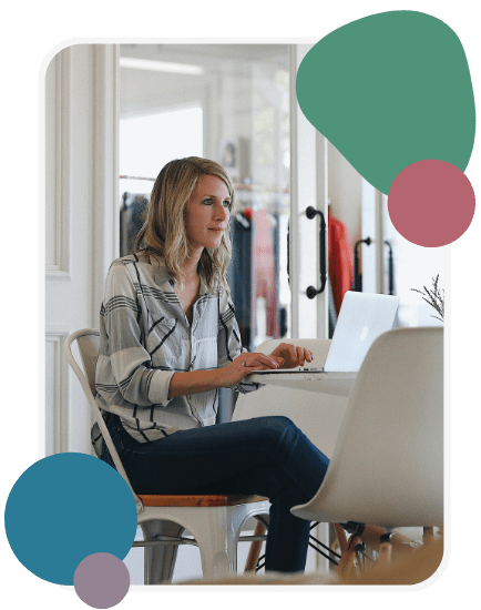 A woman with blonde hair sits at a table working on a laptop in a bright room. She wears a patterned blouse and jeans. Abstract green, blue, and maroon shapes decorate the image corners.