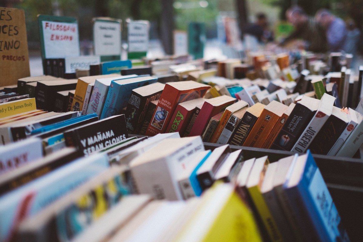 Rows of assorted books displayed on tables at an outdoor book market, with blurred signs and people in the background. Some book covers and titles are visible, creating a colourful and lively atmosphere.