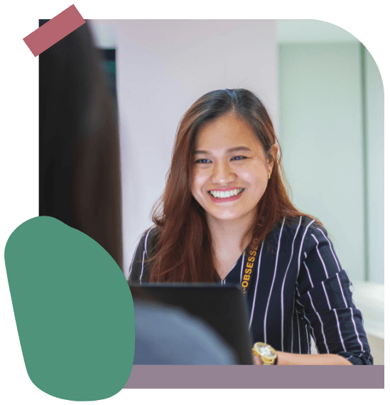 A woman with long hair and a striped shirt smiles while sitting at a desk, facing another person, in a brightly lit indoor setting.