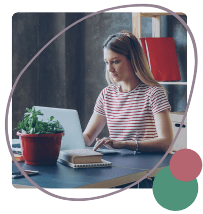 A woman in a striped shirt works on a laptop at a desk with a notebook and a potted plant.