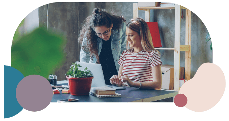 Two women smiling and collaborating at a desk with a laptop, surrounded by notebooks and a potted plant, conveying a positive, creative atmosphere.