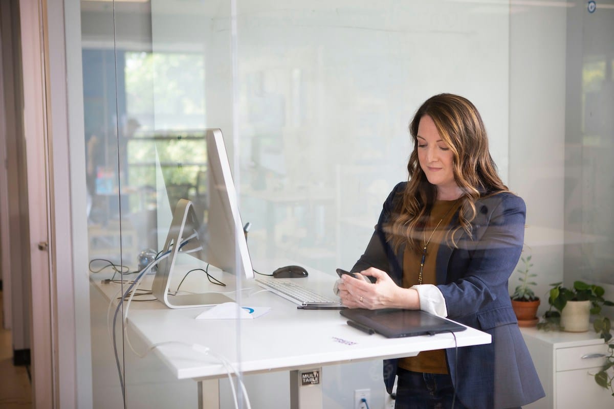 Woman in a blue blazer stands at a desk in a glass-walled office, typing on her phone. A computer and potted plants are on the desk, exuding a focused, professional atmosphere.