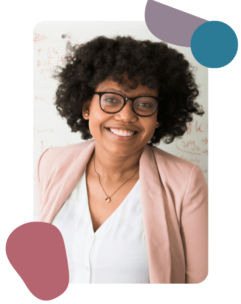 Smiling woman with curly hair and glasses, wearing a light pink blazer and white top, stands against a whiteboard backdrop. The mood is friendly and professional.