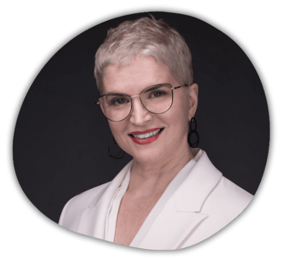A person with short, light hair wearing glasses, a white blazer, and hoop earrings smiling against a dark background.