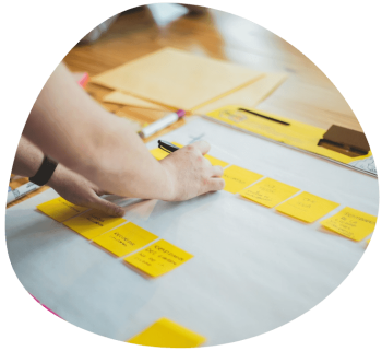 A person writes on yellow sticky notes arranged in a row on a large sheet of paper, surrounded by office stationery on a wooden table.